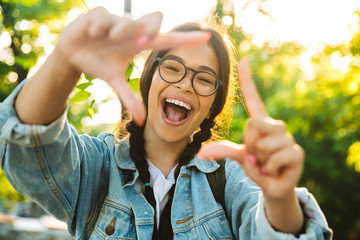 Cheerful smiling cute young student girl wearing eyeglasses outdoors in nature park make photo frame gesure.