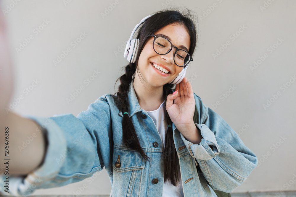 Canvas Prints Happy beautiful girl in denim jacket wearing eyeglasses isolated over gray background listening music with headphones.