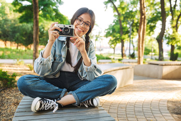 Happy cute young student girl wearing eyeglasses sitting on bench outdoors in nature park holding camera photographing.