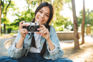 Happy cute young student girl wearing eyeglasses sitting on bench outdoors in nature park holding camera photographing.