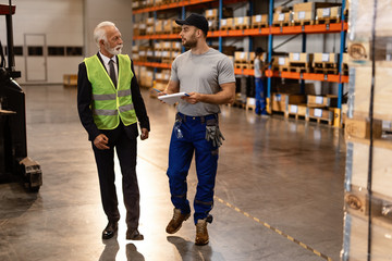 Warehouse worker and mature businessman talking while going through paperwork in industrial building.