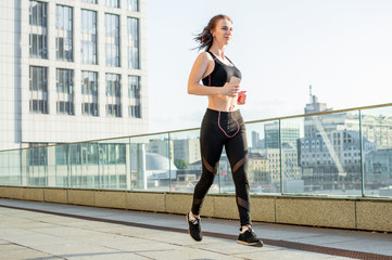 Naklejka premium athletic girl runs to workout in the morning against the background of the city and listens to music, she smiles and trains