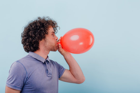 young man inflates a red balloon. with curly hair, isolated blue background, positive facial emotions, copy space