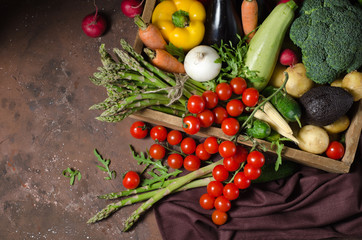 Box with farm vegetables on a dark background. Place for text. Cherry tomatoes, asparagus, broccoli, peppers, eggplants, zucchini, carrots and onions.