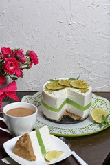 Lime cheesecake without baking. The piece is cut off. Lies on a plate. Near a cup of coffee with milk.  Against the background of white wall with decorative plaster.