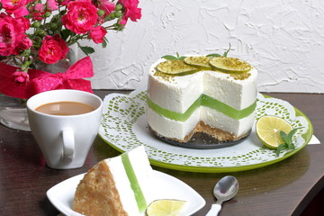 Lime cheesecake without baking. The piece is cut off. Lies on a plate. Near a cup of coffee with milk.  Against the background of white wall with decorative plaster.