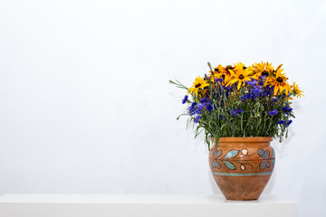 Clay vase with authentic ornament with blue and orange flowers stands on the white console opposite the white wall