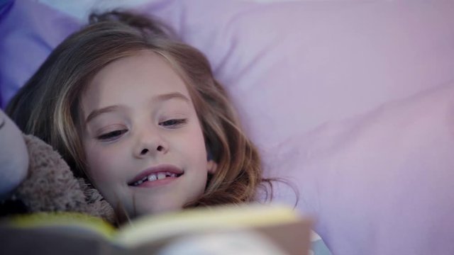 cute preteen child in pajamas lying on bed with teddy bear and smiling while reading book