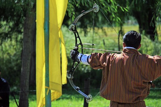 Bogensch&uuml;tze beim Training im K&ouml;nigreich Bhutan, Himalaya, Asien. Mit modernen Gelenkbogen in traditioneller Kleidung, dem Gho, zielt er im Sonnenschein zum Schuss bereit. Olympia wartet schon.
