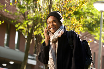 smiling young black man walking in city with mobile phone and bag during autumn
