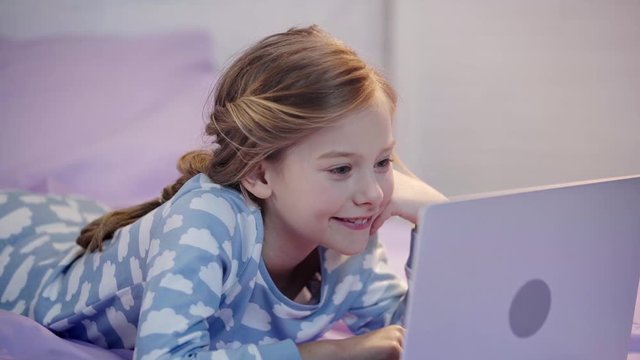 Preteen Kid In Pajamas Lying On Bed, Using Laptop And Propping Face With Hand With Smile