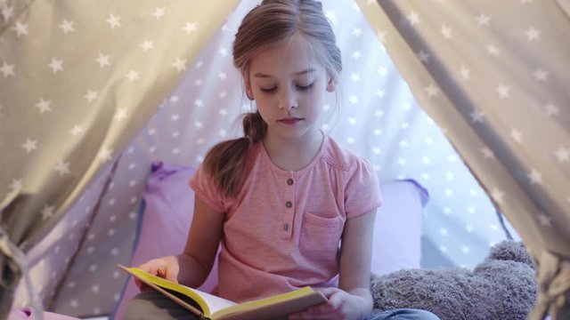 cute focused preteen kid sitting in wigwam and reading book