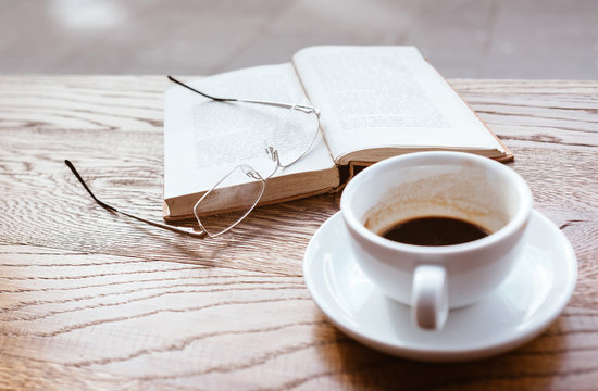 Still life image of cup of black coffee on saucer with vintage book and eyeglasses on the wooden table next to big coffee shop window.