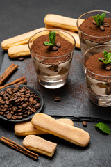 Classic tiramisu dessert in a glass and savoiardi cookies on stone serving board on dark concrete background