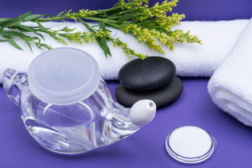 Spa purple background with Neti Pot, pile of Saline, rolled up White Towels and stacked Basalt Stones. Sinus wash. Nasal irrigation.