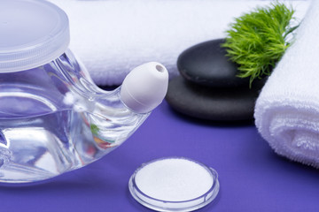 Spa purple background with Neti Pot, pile of Saline, rolled up White Towels and stacked Basalt Stones. Sinus wash. Nasal irrigation.