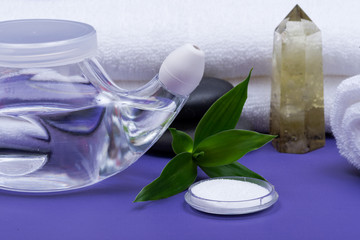 Spa purple background with Neti Pot, pile of Saline, rolled up White Towels, stacked Basalt Stones and Bamboo Leaves. Sinus wash. Nasal irrigation.