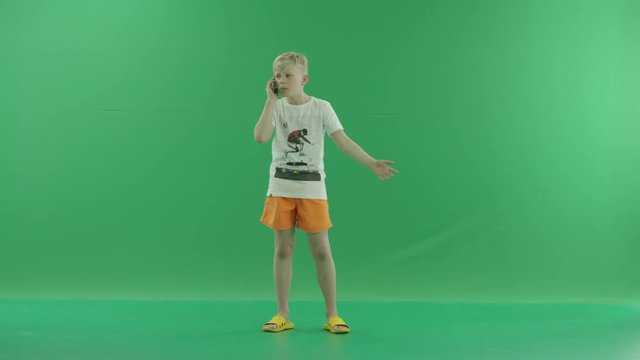 Angry young blonde boy in casual clothes is talking on his mobile phone. He stands sideways against a greenscreen