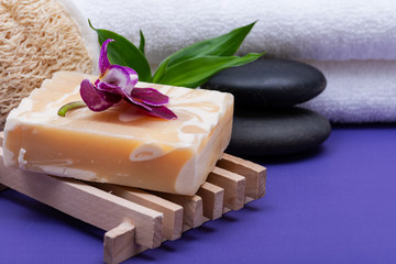 Obraz na płótnie Canvas Spa Wellness Concept. Natural Loofah Sponge, Almond Goat milk Soap on Wooden Soap Holder, Basalt Stones, Bamboo and Orchid on purple background.