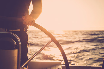 Close up of man's hand on sail boat helm - marine ship lifestyle concept of travel for beautiful holiday destination - alternative people life - sunset and sunlight in background on the ocean - obrazy, fototapety, plakaty