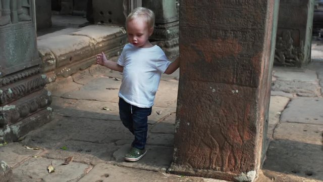 Adorable caucasian two-years old boy is running around column with carvings in Bayon temple, Angkor Wat complex, Cambodia
