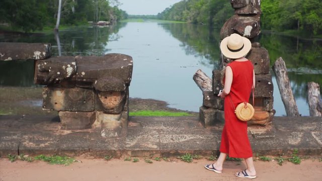 Female tourist is walking along the row of ruined asuras and devas statues exploring them. Bridge to Angkor Thom temple in Cambodia. River and jungle are on the background