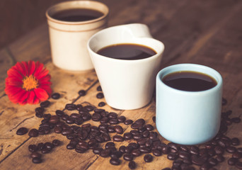Black coffee In the cup sky blue color 1 and white 2 Have all 3, cups have roasted coffee beans are on table with, still have red flowers all placed on a wooden table.