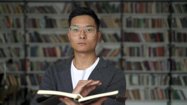 Asian man holding a blocknote swiping in the air in virtual screen with a hand turning over a page on background of bookshelves. Concept of virtual book swiping