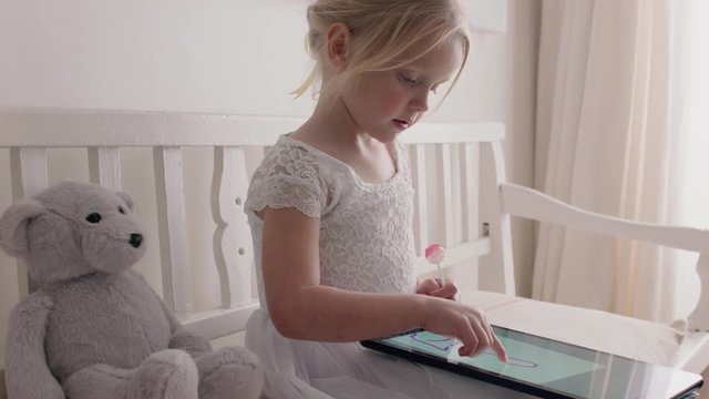 beautiful little girl using tablet computer drawing pictures on touchscreen enjoying creativity sitting on bench with teddy bear eating lollipop at home 