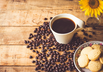 Black coffee In the cup white have roasted coffee beans dispersedly are on table, have milk cookies many pieces In the small dish, and still have sunflower, all are placed on a wooden table.