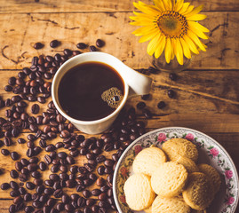 Black coffee In the cup white have roasted coffee beans dispersedly are on table, have milk cookies many pieces In the small dish, and still have sunflower, all are placed on a wooden table.
