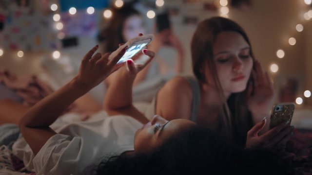 happy teenage girls lying on bed using smartphones browsing social media texting sharing gossip enjoying sleep over on weekend