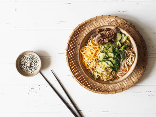 Cold Korean kuksi soup with vegetables, scrambled eggs, beef and noodles in a bowl and chopsticks on a gray background. Top view