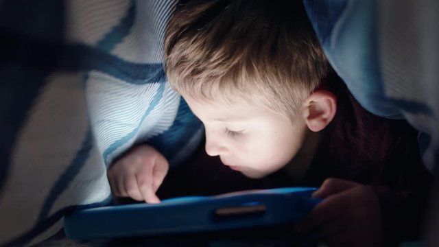 happy little boy using digital tablet computer under blanket enjoying learning on touchscreen technology playing games having fun at bedtime