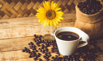 Black coffee In the cup white have roasted coffee beans In a cloth bag sack and dispersedly are on table, and still have sunflower, all are placed on a wooden table.