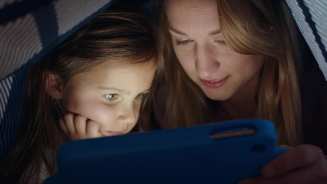 happy mother and daughter using tablet computer under blanket playing games on touchscreen technology relaxing having fun before bedtime 