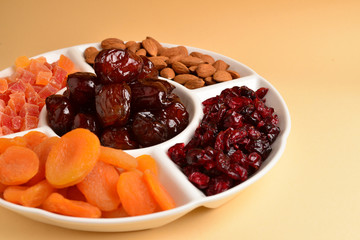 Mix of dried fruits and nuts on a white plate. Apricot, almond, raisin, dates fruit. On a brown background. Space for text or design.