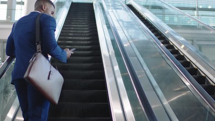 young african american businessman using smartphone on escalator texting checking email messages online successful male executive arriving at work in corporate office building 4k - Powered by Adobe
