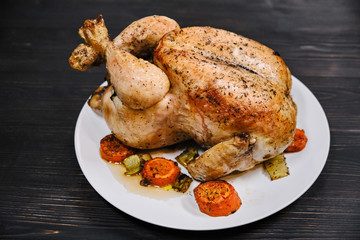 Fried with carrot chicken on black wooden background. Festive dinner with baked bird. Big whole grilled juicy chicken with golden crust on a plate.
