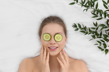 Beautiful woman with cucumber slices and leaves on white fabric, above view. Organic face mask