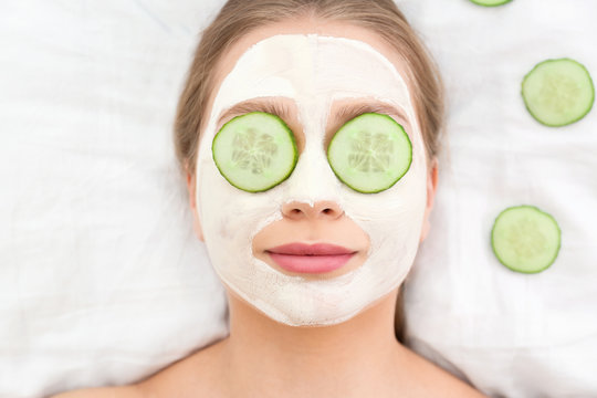 Beautiful woman with organic facial mask and cucumber slices on white fabric, above view