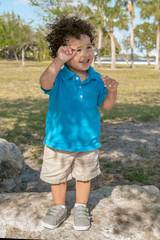 A toddler stands up on a boulder at the community park. The summer sun cascades on him as he tries to cover his eyes and smiles.