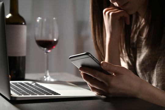 Woman using smartphone at table with laptop near glass of wine indoors, closeup. Loneliness concept