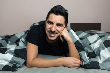 Handsome young brunette man is lying in bed, smiling and looking at the camera.