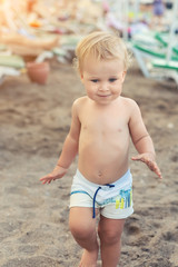 Cute caucasian toodler boy walking alone on sandy beach between chaise-lounge. Adorable happy child having fun playing at seaside shore during vacation trip
