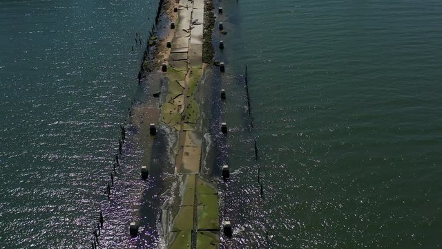 drone shot while tilting down on an old moldy, partly sunken, abandoned pier, with the sun shinning & shimmering onto the waters below. The water retaking a man-made object