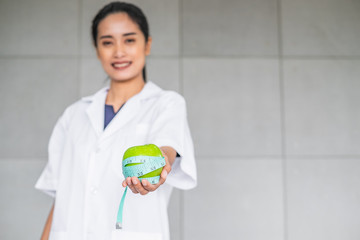 Smiling Asian woman doctor in medical white gown holding a green apple wrap with measuring tape. Health concept.