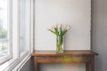 Pale pink tulips in glass vase on wooden sidetable against white painted brick wall and window with natural sun flare (selective focus)