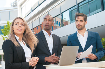 Business team with laptop watching exciting scene outdoors. Two men and woman in office suits standing outside and looking into distance. Vision concept