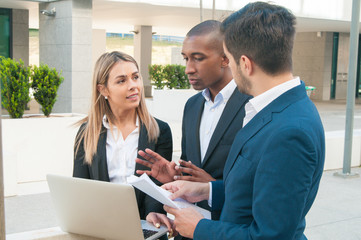 Fototapeta premium Focused business colleagues arguing while collaborating on project. Men and woman in office suits standing outdoors with laptop and papers and talking, male coworker gesturing. Cooperation concept